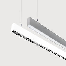 The picture shows a section of two linear pendant luminaires, one with a white and one with a grey surface. The underside of the white luminaire is fitted with a silver-coloured lenticular louvre, with each lens containing a small LED light. The grey luminaire has a smooth cover. Both luminaires have a modern design and are suspended from the ceiling on thin cables. The background is grey.