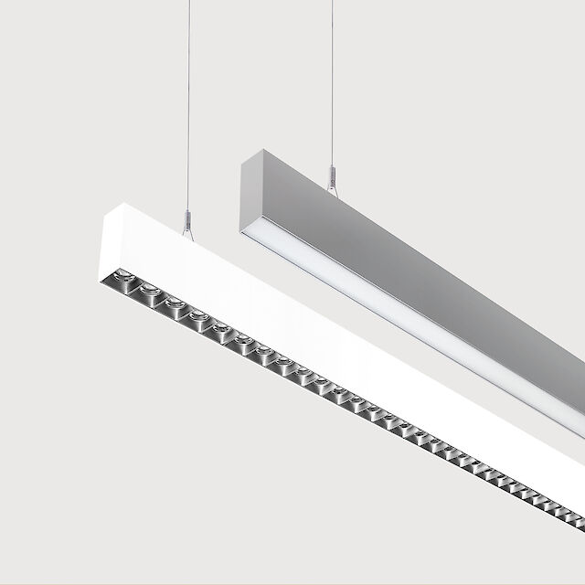 The picture shows a section of two linear pendant luminaires, one with a white and one with a grey surface. The underside of the white luminaire is fitted with a silver-coloured lenticular louvre, with each lens containing a small LED light. The grey luminaire has a smooth cover. Both luminaires have a modern design and are suspended from the ceiling on thin cables. The background is grey.