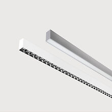 The picture shows a section of two linear surface-mounted luminaires, one with a white and one with a grey surface. The underside of the white luminaire is fitted with a black louvre, with each lens containing a small LED light. The grey luminaire has a smooth cover. Both luminaires have a modern design. The background is grey.