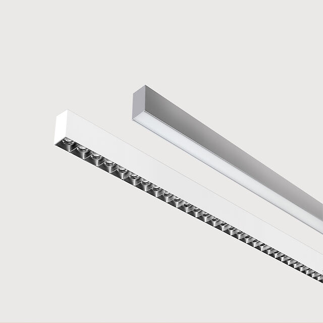 Das Bild zeigt einen Ausschnitt von zwei linearen Anbauleuchten, eine mit weißer und eine mit grauer Oberfläche. Die Unterseite der weißen Leuchte ist mit einem schwarzen Linsenraster ausgestattet, wobei jede Linse eine kleine LED-Leuchte enthält. Die graue Leuchte hat eine glatte Abdeckung. Beide Leuchten sind modern gestaltet. Der Hintergrund ist grau.