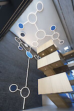 Innenansicht eines modernen Gebäudes mit einer auffälligen Beleuchtungsinstallation an der Decke, bestehend aus großen, leuchtenden Kreisen, die durch Linien verbunden sind, ähnlich wie Moleküle. Einige Kreise sind blau. Die Wände sind aus dunklen Ziegeln und kontrastieren mit hellen, hölzernen Treppen und Plattformen über mehrere Etagen. Das Licht und die Materialien schaffen eine offene, futuristische Atmosphäre.