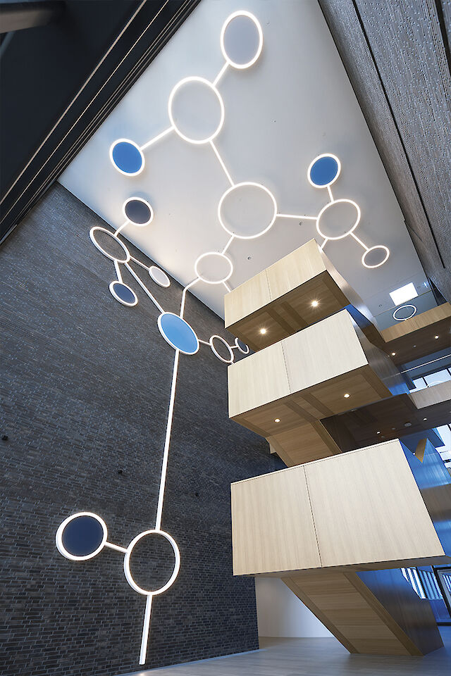 An eye-catching light installation on the ceiling inside a modern building, consisting of large, luminous circles connected by lines, similar to molecules. Some of the circles are blue. The walls are made of dark bricks and contrast with light-coloured, wooden stairs and platforms over several floors. The light and materials create an open, futuristic atmosphere.