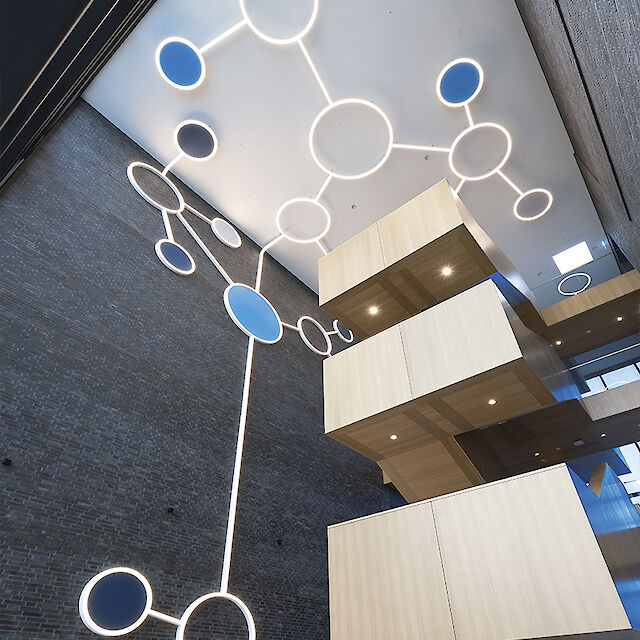 An eye-catching light installation on the ceiling inside a modern building, consisting of large, luminous circles connected by lines, similar to molecules. Some of the circles are blue. The walls are made of dark bricks and contrast with light-coloured, wooden stairs and platforms over several floors. The light and materials create an open, futuristic atmosphere.