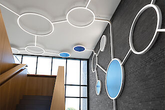 Innenansicht eines modernen Treppenhauses mit einer auffälligen Lichtinstallation an Wänden und Decke, die aus großen leuchtenden Kreisen besteht, die durch Linien verbunden sind, die an Moleküle erinnern. Einige Kreise sind blau. Die Wände sind aus dunklem Backstein und kontrastieren mit der hellen Holztreppe. Licht und Materialien schaffen eine offene, futuristische Atmosphäre.