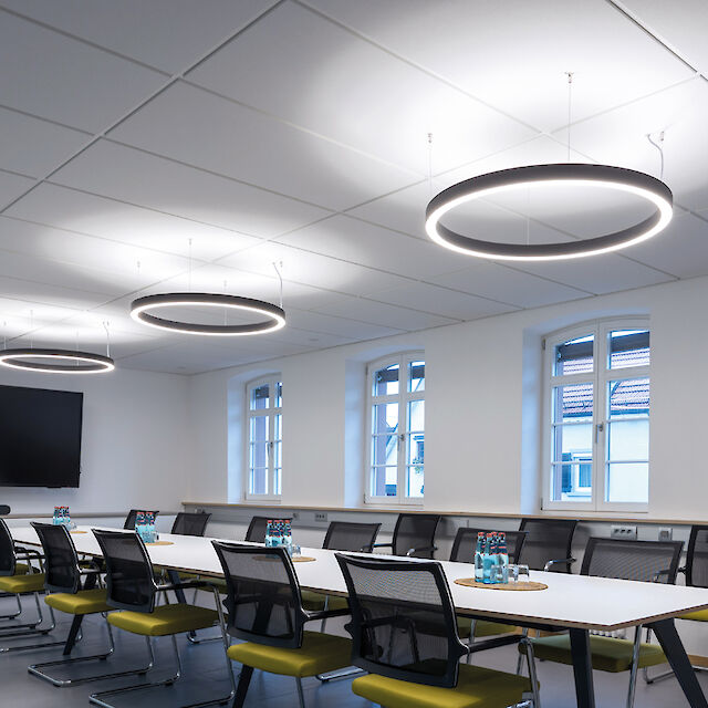 La imagen muestra una moderna sala de conferencias con largas mesas blancas y sillas negras tapizadas con respaldos de rejilla. Del techo cuelgan luces redondas en forma de anillo que iluminan la sala de manera uniforme. En la pared de la izquierda hay una gran pantalla plana. Hay varias ventanas por las que entra luz natural. Hay varias botellas de agua y vasos sobre las mesas, lo que indica que se va a celebrar una reunión. El ambiente es luminoso y acogedor.