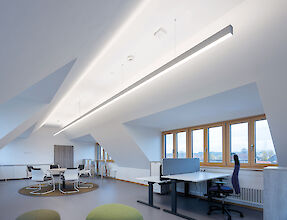 La imagen muestra una oficina moderna con techos inclinados y grandes ventanales por los que entra mucha luz natural. La sala está dividida en dos zonas: En primer plano hay dos mesas con sillas ergonómicas y monitores diseñados para el trabajo concentrado. Al fondo, una mesa redonda con varias sillas blancas. La iluminación corre a cargo de una larga y delgada luminaria en el techo, que ilumina la sala de forma brillante y uniforme. El suelo está cubierto de moqueta gris y hay una alfombra redonda cerca del rincón de reunión. El ambiente general es abierto y acogedor.