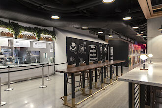 Vista interior del Moynihan Food Hall con puesto de autoservicio, mesas altas y luminarias suspendidas Beam Me Up.