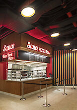 Vista interior del Moynihan Food Hall: puesto de pizzas con mostrador de cocina abierto y luminarias suspendidas Beam Me Up.