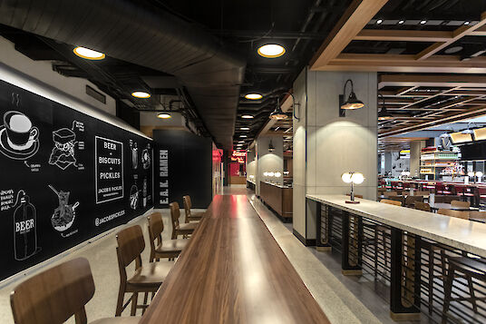 Vista interior del Moynihan Food Hall con largas mesas de madera, gráficos murales y luminarias suspendidas Beam Me Up.