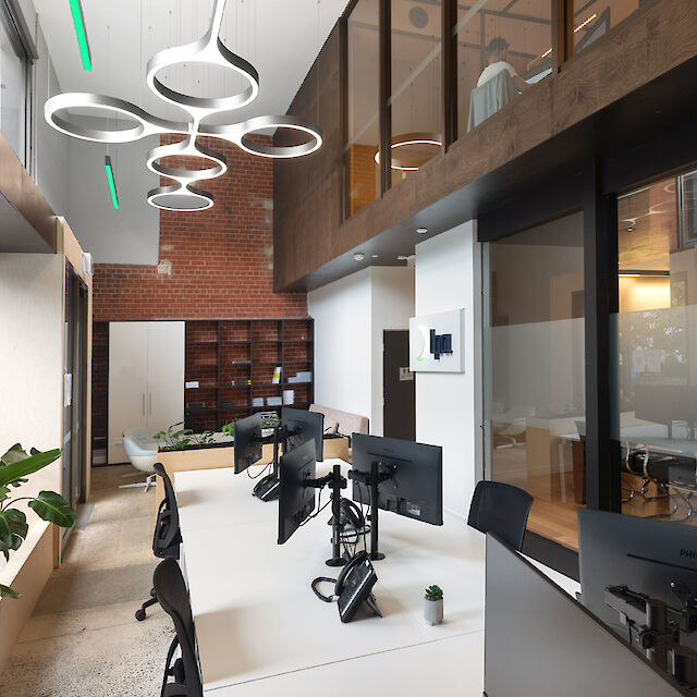 A modern office with high ceilings and an open-plan layout. There are large windows on the left that let in plenty of daylight. Plants hang on the walls, adding a green touch. In the centre of the room are desks with monitors and office chairs. Other offices and shelves are visible in the background. The ceiling is decorated with stylish, round pendant luminaires. The floor is made of a light-coloured material that gives the room a bright and inviting ambience.
