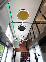 Un moderno edificio de oficinas con techo alto. Del techo cuelgan varias luminarias circulares modernas con diferentes elementos acústicos de distintos colores (amarillo, negro) y formas. A la izquierda hay grandes ventanales por los que entra la luz del día, así como una línea de luz verde resplandeciente en el techo. A la derecha hay oficinas acristaladas en la planta superior. Al fondo se ve una pared de ladrillos rojos y estanterías.

