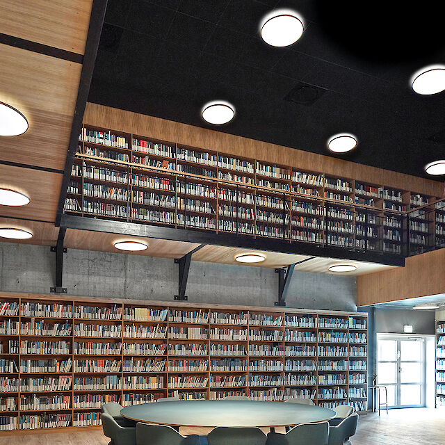 The picture shows a modern library with high bookshelves running along the walls and on an intermediate level. The bookshelves are filled with a variety of books. In the centre of the room is a round table with several chairs that invite you to read and work. The ceiling is black and fitted with several round, copper-coloured surface mounted luminaires that ensure the room is evenly lit. The walls and shelves are made of light-coloured wood, which gives the room a warm and inviting atmosphere. A large entrance with glass doors can be seen on the right-hand side of the picture, which lets in plenty of natural light.



