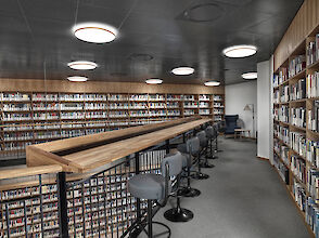 The picture shows an interior view of the university library in Aalborg, Denmark. The room is furnished with high bookshelves that fill the walls and house a large number of books. In the foreground of the picture is a long wooden counter with several modern, upholstered high chairs that have black metal frames and round seats and backrests. The ceiling is fitted with several round, copper-coloured surface-mounted luminaires that provide even lighting in the room. The atmosphere is calm and inviting, ideal for reading and studying.