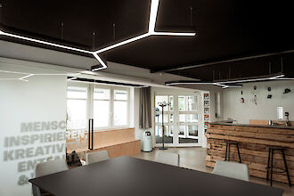 The picture shows a modern room with a mixture of office and lounge areas. Eye-catching, geometrically shaped LED luminaires are mounted on the ceiling. In the foreground is a large black table with several light grey chairs. The words 