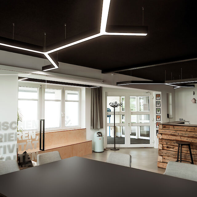 The picture shows a modern room with a mixture of office and lounge areas. Eye-catching, geometrically shaped LED luminaires are mounted on the ceiling. In the foreground is a large black table with several light grey chairs. The words 