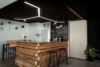 The picture shows a modern café or bar corner with a rustic wooden bar counter. Above the counter, geometrically shaped LED luminaires hang from the dark ceiling. A door leading to another room can be seen in the background. The wall behind the counter is covered with a blackboard on which handwritten menu items can be seen. There are two black bar stools in front of the counter.