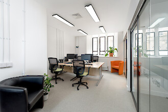 Bureau équipé de tables, d'ordinateurs, de chaises pivotantes, de plantes et de luminaires linéaires au plafond, dans une pièce lumineuse
