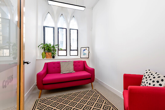 Petite salle de réunion avec un canapé rouge, un fauteuil, une plante et une suspension linéaire devant des fenêtres en ogive