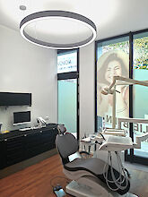 Treatment room of a dental practice with ring-shaped LED pendant luminaires and dentist's chair in front of a large window front.