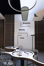 The picture shows a modern, stylish room with several curved tables and chairs. Three different pendant lights hang from the white ceiling. The names 