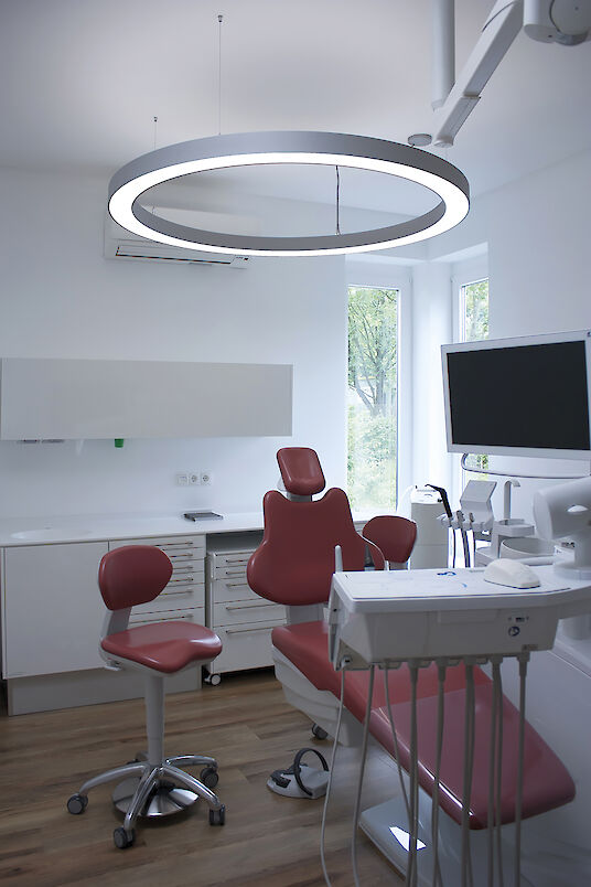 Cabinet dentaire avec fauteuil de soins rouge, écran et luminaire suspendu Ringo Star en forme d'anneau.