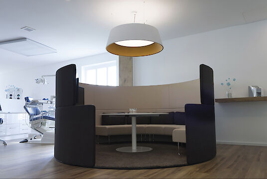 Espace lounge dans un cabinet dentaire avec une niche ronde et un grand luminaire suspendu Beam Me Up avec réflecteur doré.