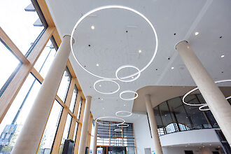 Habitación diáfana y luminosa con techos altos y grandes ventanales que dejan pasar la luz natural. Varias luminarias colgantes en forma de anillo de distintos tamaños cuelgan del techo y crean un concepto de iluminación moderno y minimalista. Unas columnas macizas y una pared suavemente curvada dan a la estancia estructura y profundidad arquitectónica.