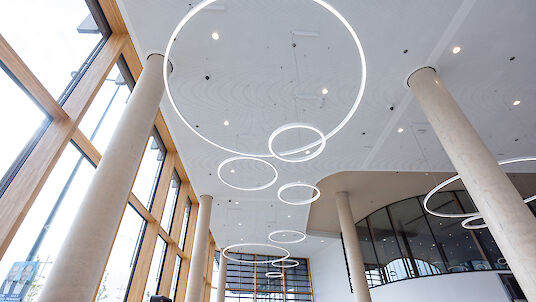 Bright, open room with a high ceiling and large windows that let in natural light. Several ring-shaped pendant luminaires in different sizes hang from the ceiling and create a modern and minimalist lighting concept. Massive columns and a gently curved wall give the room structure and architectural depth.