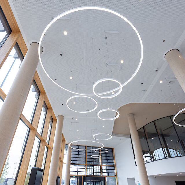 Bright, open room with a high ceiling and large windows that let in natural light. Several ring-shaped pendant luminaires in different sizes hang from the ceiling and create a modern and minimalist lighting concept. Massive columns and a gently curved wall give the room structure and architectural depth.
