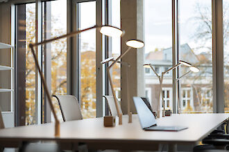 Modern office space with large floor-to-ceiling windows that let in plenty of natural light and offer a view of the autumnal outdoors. On a long desk are several minimalist, white desk luminaire with a modern look. A laptop is open on the desk and white office chairs with mesh-like backrests are arranged around the table. White shelves can be seen in the background and the walls and ceilings have an industrial concrete look.