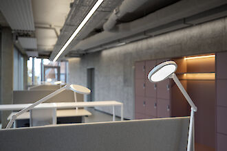 The picture shows a modern office with a row of workstations separated by grey partitions. In the foreground is a white desk luminaire with a round, flat luminaire head. The ceiling has an industrial design with visible pipes and cable ducts. A long, narrow line of light runs along the ceiling, illuminating the room evenly. Grey cabinets with integrated shelves and lockers can be seen on the right-hand side of the picture. 