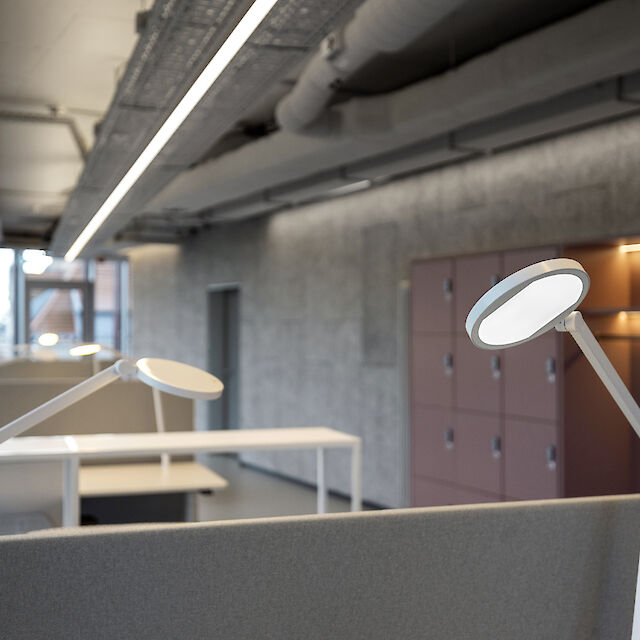 The picture shows a modern office with a row of workstations separated by grey partitions. In the foreground is a white desk luminaire with a round, flat luminaire head. The ceiling has an industrial design with visible pipes and cable ducts. A long, narrow line of light runs along the ceiling, illuminating the room evenly. Grey cabinets with integrated shelves and lockers can be seen on the right-hand side of the picture. 