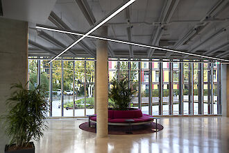 Modern, light-flooded reception area with large floor-to-ceiling windows that offer a view of the outside of a building complex with colourful facades. In the centre of the room is a circular seating area with a semi-circular, magenta-coloured couch grouped around a large plant in a planter. The floor is made of polished, shiny material. The ceiling has an industrial design with exposed beams and cables, along which linear LED strip luminaires run in geometric patterns, illuminating the space evenly. 