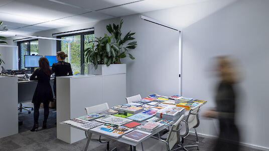 In einem modernen Büro arbeiten zwei Personen an ihren Schreibtischen, die mit Monitoren und Bürozubehör ausgestattet sind. Im Vordergrund befindet sich ein Tisch, der mit einer Vielzahl von Zeitschriften und Broschüren bedeckt ist. Pflanzen verleihen dem Raum eine grüne Note. Die Beleuchtung ist hell und gleichmäßig, unterstützt durch schlichte, weiße Deckenleuchten. Eine Person ist unscharf im Bild, in Bewegung an der rechten Seite des Raumes.