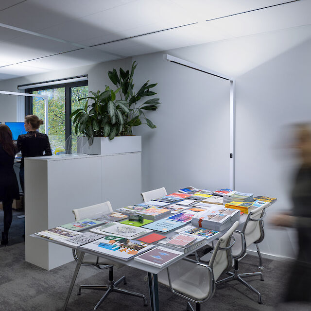 In einem modernen Büro arbeiten zwei Personen an ihren Schreibtischen, die mit Monitoren und Bürozubehör ausgestattet sind. Im Vordergrund befindet sich ein Tisch, der mit einer Vielzahl von Zeitschriften und Broschüren bedeckt ist. Pflanzen verleihen dem Raum eine grüne Note. Die Beleuchtung ist hell und gleichmäßig, unterstützt durch schlichte, weiße Deckenleuchten. Eine Person ist unscharf im Bild, in Bewegung an der rechten Seite des Raumes.