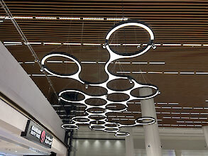 Moderne, ringförmige Leuchten von Lightnet, die an der Decke des Bahrain Duty Free Shops im Bahrain International Airport hängen. Die Leuchten sind kreisförmig angeordnet und sorgen für eine gleichmäßige, blendfreie Beleuchtung. Im Hintergrund sind die Holzelemente der Decke und das Schild des Bahrain Duty Free Shops zu sehen.