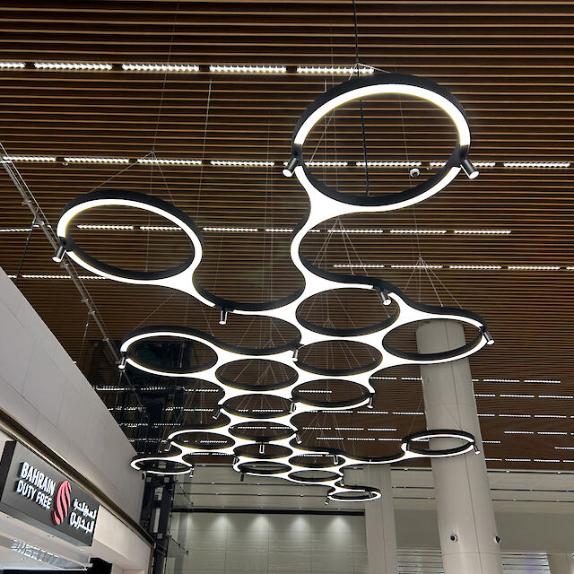 Modern, ring-shaped luminaires from Lightnet suspended from the ceiling of the Bahrain Duty Free Shop at Bahrain International Airport. The luminaires are arranged in a circle and provide uniform, glare-free lighting. The wooden elements of the ceiling and the Bahrain Duty Free Shop sign can be seen in the background.