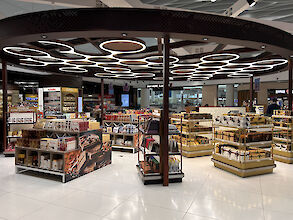 Innenansicht des Bahrain Duty Free Shops im Bahrain International Airport, beleuchtet von ringförmigen Ringo Star Cluster-G3 Leuchten an der Decke. Die Verkaufsfläche ist mit Regalen gefüllt, die verschiedene Süßwaren und Schokoladenprodukte präsentieren. Die Beleuchtung sorgt für eine gleichmäßige und angenehme Ausleuchtung des Bereichs. Im Hintergrund sind weitere Verkaufsstände und Kunden zu sehen.