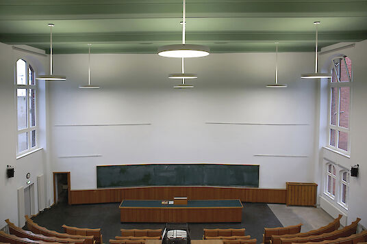 Lecture theatre with green ceiling, large blackboard, wooden desk and round Basic pendant luminaires.