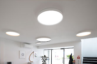 Un techo con varias luminarias redondas y planas distribuidas uniformemente. Las luminarias emiten una luz brillante y agradable que ilumina bien la estancia. A continuación, una zona de oficina moderna y luminosa con obras de arte en las paredes, una planta de interior y una gran ventana que deja pasar la luz natural.