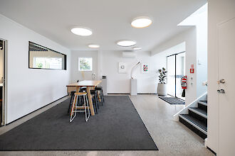 Un espacio de oficina moderno con varias lámparas de techo redondas que emiten una luz brillante y agradable. En el centro de la sala hay una larga mesa de madera con varios taburetes blancos y negros. Dos obras de arte enmarcadas cuelgan de la pared. Una gran ventana y una puerta de cristal dejan pasar la luz natural. A la derecha hay una escalera que sube y, junto a ella, una gran planta de interior en una maceta blanca. La estancia resulta abierta y acogedora.