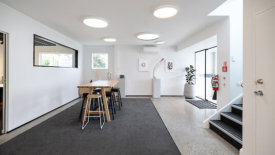 A modern office space with several round ceiling lights that emit a bright, pleasant light. In the centre of the room is a long wooden table with several black and white stools. Two framed works of art hang on the wall. A large window and a glass door let in natural light. On the right-hand side, a staircase leads upwards and next to it is a large houseplant in a white pot. The room feels open and inviting.