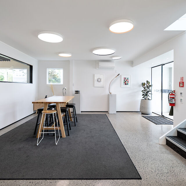 A modern office space with several round ceiling lights that emit a bright, pleasant light. In the centre of the room is a long wooden table with several black and white stools. Two framed works of art hang on the wall. A large window and a glass door let in natural light. On the right-hand side, a staircase leads upwards and next to it is a large houseplant in a white pot. The room feels open and inviting.