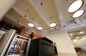 The picture shows a modern reception area in a building. In the foreground is a black reception counter with a person standing behind it. On the wall behind the counter is a wooden panelling with the inscription 