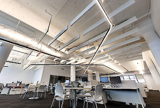 The picture shows a modern, open-plan office with high ceilings. The ceiling is fitted with exposed ventilation ducts and white acoustic panels. A striking, curved LED light line runs through the room and provides illumination. The workstation area consists of white desks, ergonomic office chairs and several monitors. In the foreground are a few tables and chairs for meetings or collaboration. The room feels bright and spacious.