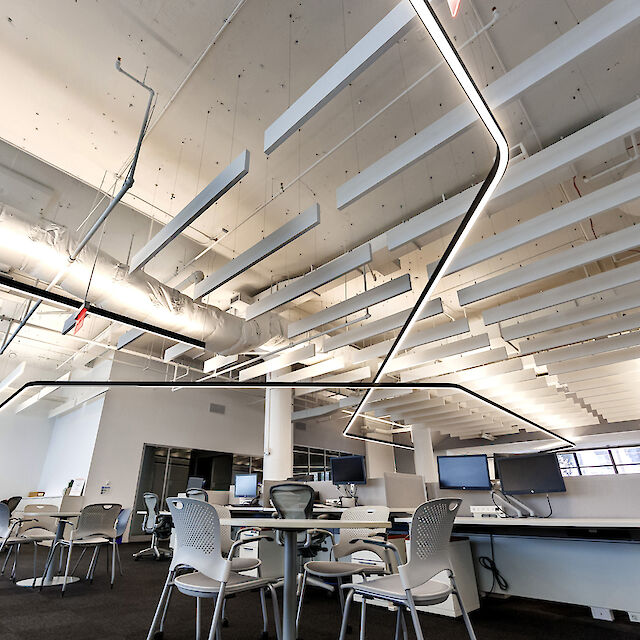 La imagen muestra una moderna oficina diáfana con techos altos. El techo está equipado con conductos de ventilación a la vista y paneles acústicos blancos. Una llamativa línea de luz LED curvada recorre la sala y proporciona iluminación. La zona de trabajo está formada por mesas blancas, sillas de oficina ergonómicas y varios monitores. En primer plano hay algunas mesas y sillas para reuniones o colaboración. La sala es luminosa y espaciosa.