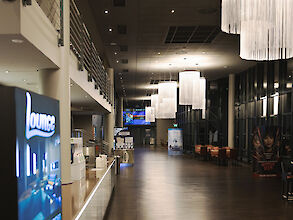 The picture shows the interior of a modern lounge in a cinema. In the foreground on the left is an illuminated sign with the inscription ‘Lounge’. The ceiling is decorated with several large, cylindrical chandeliers with long, white fringes that illuminate the room. There are several tables and chairs against the wall on the right. The back wall consists of large windows that allow a view outside. Film posters hang on the right-hand wall. In the background is a long, wide corridor with more seating and posters, creating an inviting and spacious atmosphere.