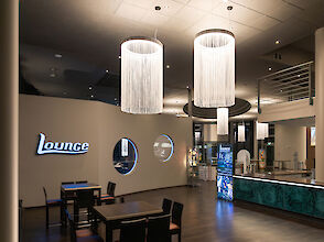 The picture shows a modern lounge with stylish furnishings. In the foreground are several black tables and chairs on a dark wooden floor. On the wall to the left is the word ‘Lounge’ in bright letters. The wall has round windows through which you can see into another area. Several large, cylindrical chandeliers with long, white fringes hang from the ceiling, illuminating the room. To the right of the picture is a counter with a green-patterned front, behind which is a digital display. Other areas of the lounge can be seen in the background, creating an inviting and open impression.