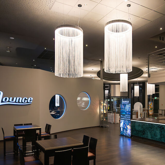 The picture shows a modern lounge with stylish furnishings. In the foreground are several black tables and chairs on a dark wooden floor. On the wall to the left is the word ‘Lounge’ in bright letters. The wall has round windows through which you can see into another area. Several large, cylindrical chandeliers with long, white fringes hang from the ceiling, illuminating the room. To the right of the picture is a counter with a green-patterned front, behind which is a digital display. Other areas of the lounge can be seen in the background, creating an inviting and open impression.