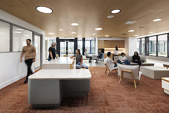 Open lounge area with wooden ceiling, round recessed lights, carpeted floor, seating and people working.